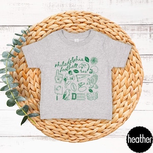 May include: A heather gray t-shirt featuring a green graphic design with the words "Philadelphia Football" and various football-related illustrations. The design includes an eagle, footballs, and a stadium.