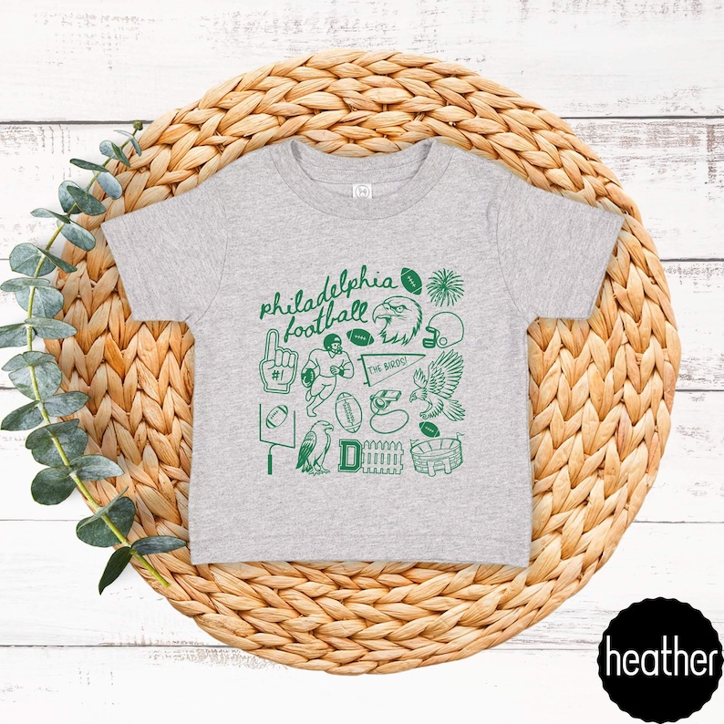 May include: A heather gray t-shirt featuring a green graphic design with the words "Philadelphia Football" and various football-related illustrations. The design includes an eagle, footballs, and a stadium.