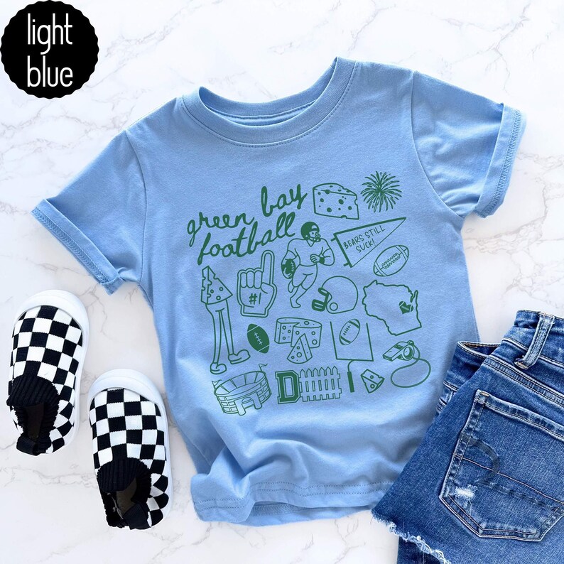 May include: Pale blue t-shirt featuring a green "Green Bay Football" design and football-related images. Includes a pair of black and white chequered slip-on shoes and denim shorts.