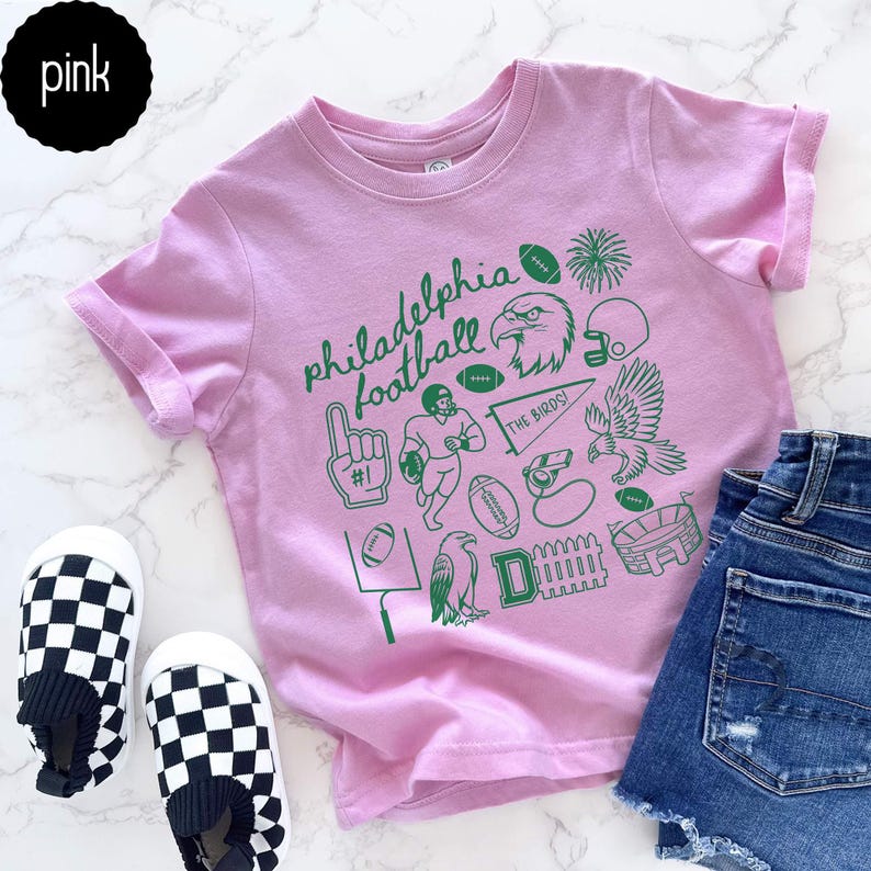 May include: A pink t-shirt with green graphics and the text "Philadelphia Football." The design includes footballs, an eagle, a helmet, and a stadium. The shirt is paired with denim shorts and checkered slip-on shoes.