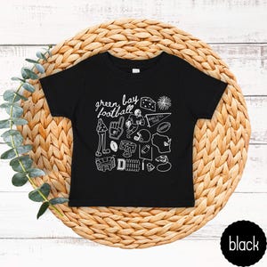 May include: Black toddler t-shirt with a white graphic design featuring the words "green bay football" and various football-related illustrations. The shirt is displayed on a woven, circular placemat.