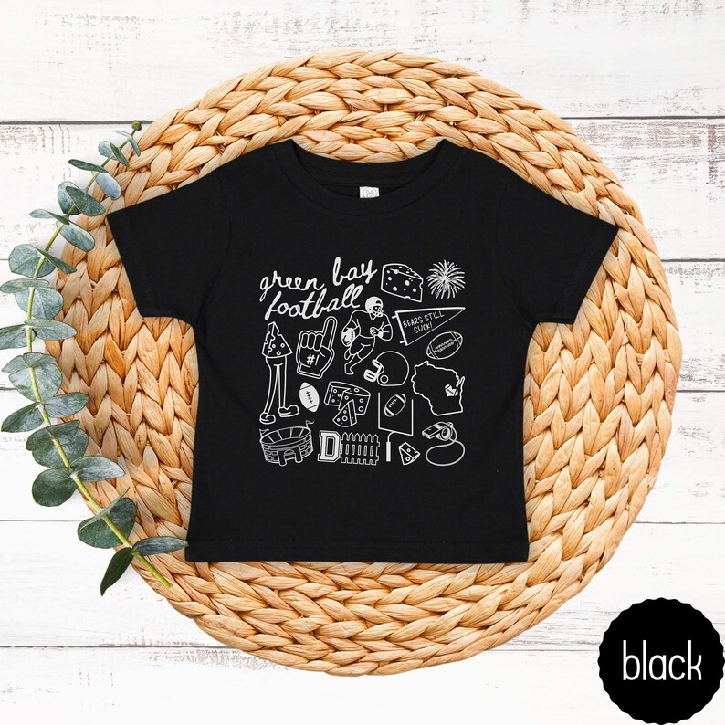 May include: Black toddler t-shirt with a white graphic design featuring the words "green bay football" and various football-related illustrations. The shirt is displayed on a woven, circular placemat.