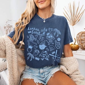 May include: Navy blue t-shirt with a light blue "Kansas City Baseball" graphic. The design features baseball-themed illustrations. The shirt is worn with denim shorts. The image shows a casual, relaxed style.