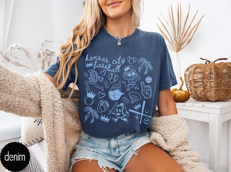 May include: Navy blue t-shirt with a light blue "Kansas City Baseball" graphic. The design features baseball-themed illustrations. The shirt is worn with denim shorts. The image shows a casual, relaxed style.