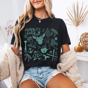 May include: Black t-shirt with a teal graphic design featuring Seattle baseball-themed illustrations. The design includes the text "Seattle baseball", a moose with sunglasses, and various baseball-related icons.