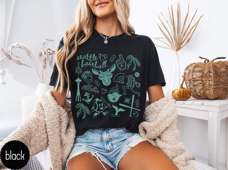 May include: Black t-shirt with a teal graphic design featuring Seattle baseball-themed illustrations. The design includes the text "Seattle baseball", a moose with sunglasses, and various baseball-related icons.