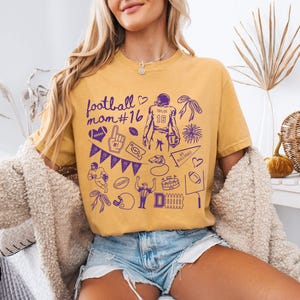 May include: A mustard yellow short-sleeve t-shirt featuring a purple graphic print with the text football mom #16, a football player, a foam finger, a whistle, a pennant, a stadium, and various football-themed icons including hearts and bows.