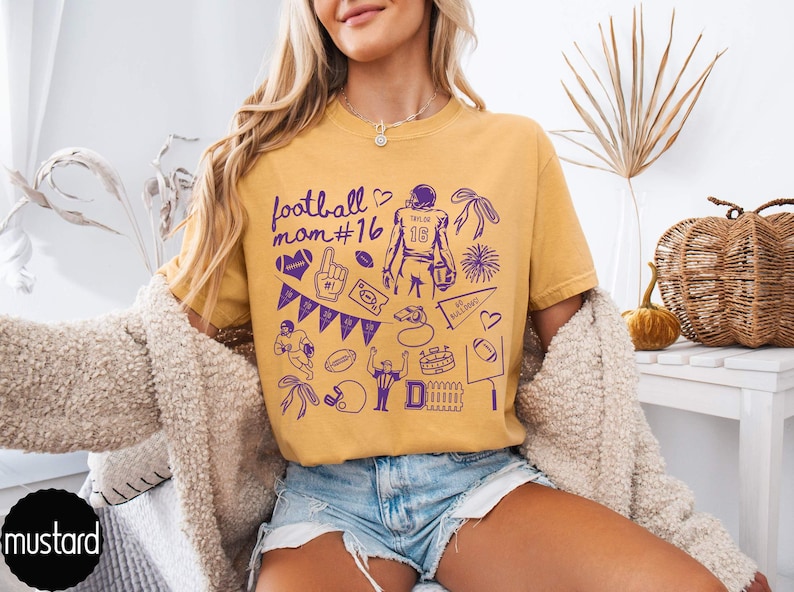 May include: A mustard yellow short-sleeve t-shirt featuring a purple graphic print with the text football mom #16, a football player, a foam finger, a whistle, a pennant, a stadium, and various football-themed icons including hearts and bows.