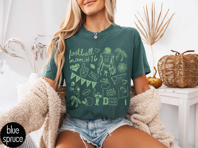 May include: A dark green short-sleeve t-shirt featuring light green line art graphics related to football, including a player, a stadium, a pennant, and the text "football mom #16". The shirt has a crew neckline and a relaxed fit.