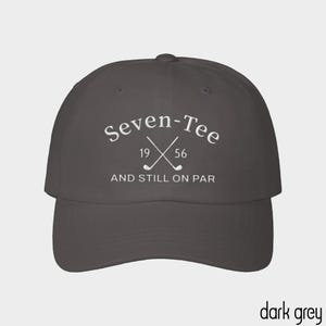 May include: A dark grey baseball cap featuring white embroidered text that reads Seven-Tee with the numbers 19 and 56 flanking two crossed golf clubs, followed by the text AND STILL ON PAR centered below the golf club graphic.