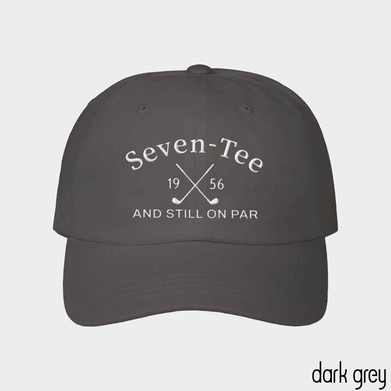 May include: A dark grey baseball cap featuring white embroidered text that reads Seven-Tee with the numbers 19 and 56 flanking two crossed golf clubs, followed by the text AND STILL ON PAR centered below the golf club graphic.
