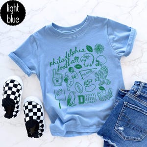 May include: Light blue t-shirt with green Philadelphia football graphics, including an eagle, footballs, and the text "Philadelphia football". The shirt is paired with checkered slip-on shoes and denim shorts.