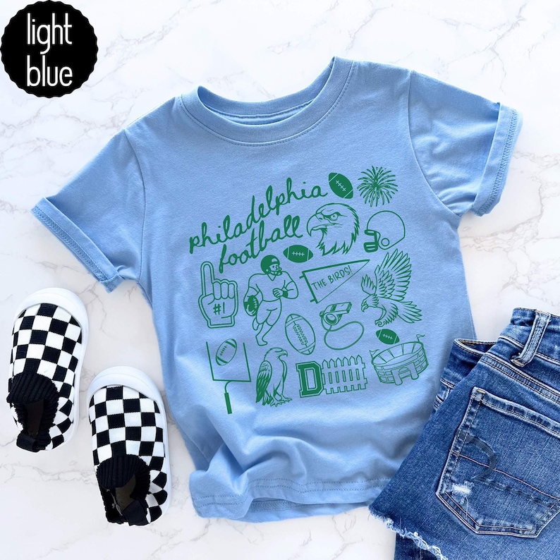 May include: Light blue t-shirt with green Philadelphia football graphics, including an eagle, footballs, and the text "Philadelphia football". The shirt is paired with checkered slip-on shoes and denim shorts.