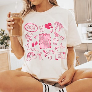 May include: Cream-colored t-shirt with pink illustrations of baby-related items, including a stroller, footprints, and a rocking chair. The shirt features the text "Olivia's Mama" and "est. 2024". The design includes a heart, bow, and a rainbow.