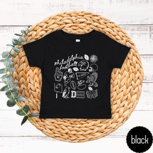 May include: Black toddler t-shirt with white graphic illustrations and the words "philadelphia football". The design includes a football, eagle, helmet, and other sports-related images. The shirt is displayed on a woven, circular mat.