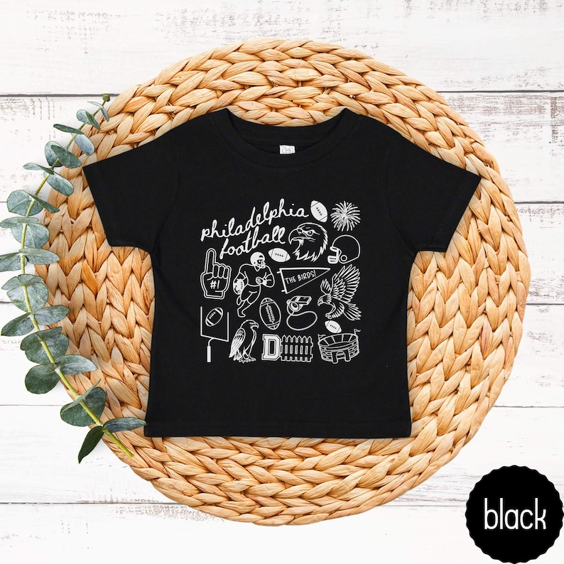 May include: Black toddler t-shirt with white graphic illustrations and the words "philadelphia football". The design includes a football, eagle, helmet, and other sports-related images. The shirt is displayed on a woven, circular mat.