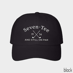 May include: A black baseball cap featuring white embroidered text that reads Seven-Tee 19 56 AND STILL ON PAR, with a graphic of two crossed golf clubs positioned between the numbers 19 and 56 on the front panel of the hat.
