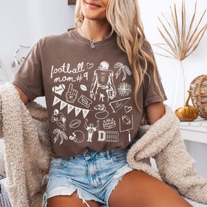 May include: A brown short-sleeve t-shirt featuring white line art graphics related to American football, including the text "football mom #9", a player, a helmet, a whistle, a pennant, a stadium, and a goalpost, paired with blue denim shorts.