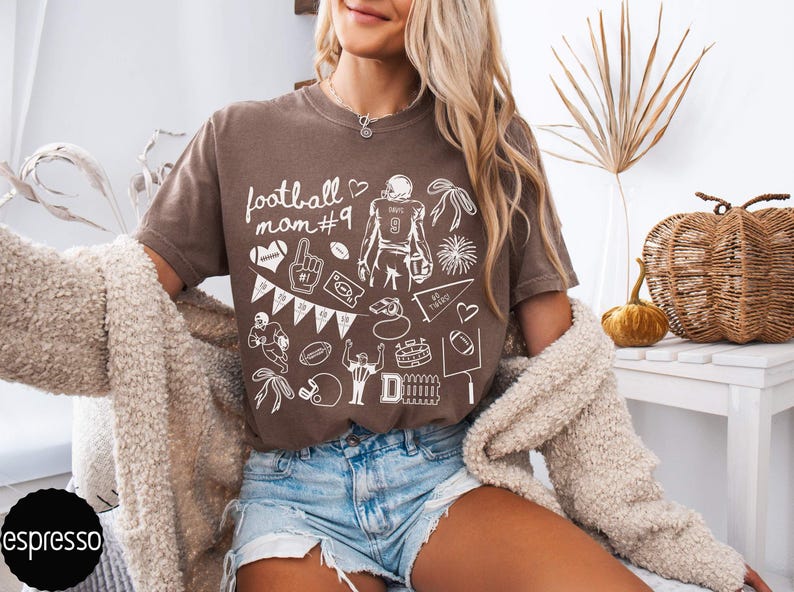 May include: A brown short-sleeve t-shirt featuring white line art graphics related to American football, including the text "football mom #9", a player, a helmet, a whistle, a pennant, a stadium, and a goalpost, paired with blue denim shorts.
