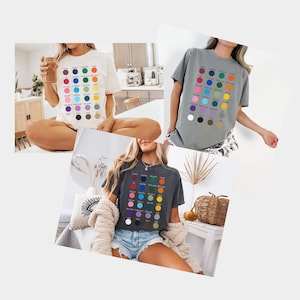 May include: Three short-sleeve t-shirts in white, gray, and charcoal, each featuring a grid of twenty-five colored circles with corresponding text labels underneath, including maroon, navy, forest green, orange, pink, light blue, lime, yellow, purple, black, brown, and gray.