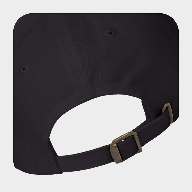 May include: A solid black baseball cap featuring a fabric strap closure at the back. The strap is secured with an antique brass-toned metal sliding buckle. The cap includes small circular stitched eyelets on the upper panels for ventilation.