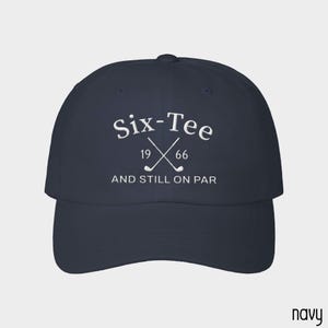 May include: A navy blue baseball cap featuring white embroidered text that reads Six-Tee 19 66 AND STILL ON PAR, with a graphic of two crossed golf clubs positioned between the numbers 19 and 66 on the front panel of the hat.