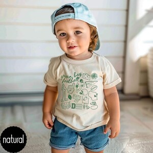 May include: A toddler wearing a pale blue baseball cap and a cream t-shirt with green illustrations and the words "green bay football". The shirt features football graphics and the word "natural" on a black circle.