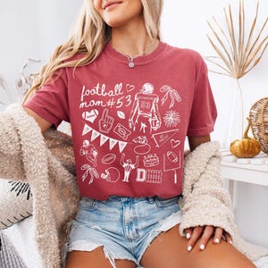 May include: A crimson short-sleeved t-shirt featuring white line art graphics related to football. The design includes the text "football mom #53", a football player, a helmet, a whistle, a pennant, a stadium, and various other sports-themed icons.
