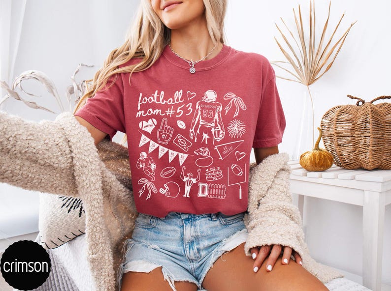 May include: A crimson short-sleeved t-shirt featuring white line art graphics related to football. The design includes the text "football mom #53", a football player, a helmet, a whistle, a pennant, a stadium, and various other sports-themed icons.