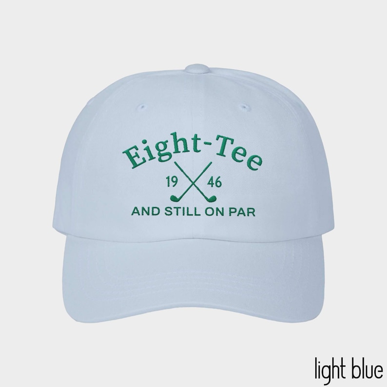 Puede incluir: Una gorra de b&eacute;isbol azul claro con bordado verde oscuro en la parte delantera. El texto dice Eight-Tee con los n&uacute;meros 19 y 46 flanqueando dos palos de golf cruzados, seguido del texto AND STILL ON PAR centrado debajo de los palos.