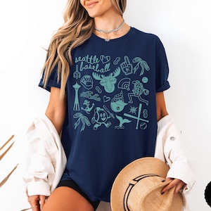May include: Navy blue t-shirt with a teal graphic design featuring Seattle-themed illustrations and the words "Seattle Baseball." The design includes a heart, moose, baseball, and other icons. The shirt is a comfortable, casual style.