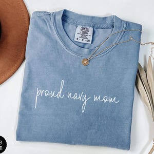 May include: A blue Comfort Colors t-shirt with the words "proud navy mom" in white script. A gold necklace with a circular pendant rests on the shirt. A brown hat and dried plant are in the background.