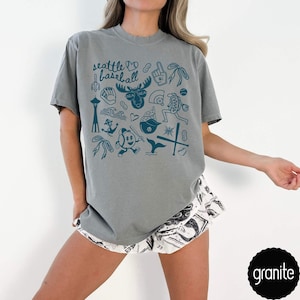 May include: A gray t-shirt with a teal graphic design featuring the text "Seattle Baseball" and various Seattle-themed illustrations. The shirt is paired with patterned shorts. The word "granite" is in a black circle.