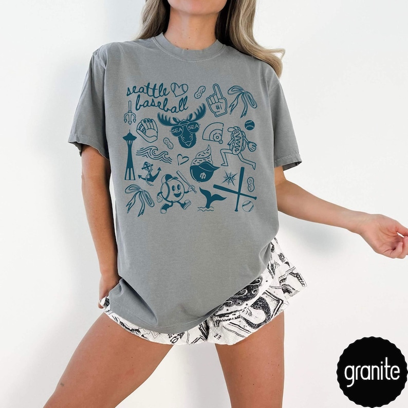 May include: A gray t-shirt with a teal graphic design featuring the text "Seattle Baseball" and various Seattle-themed illustrations. The shirt is paired with patterned shorts. The word "granite" is in a black circle.