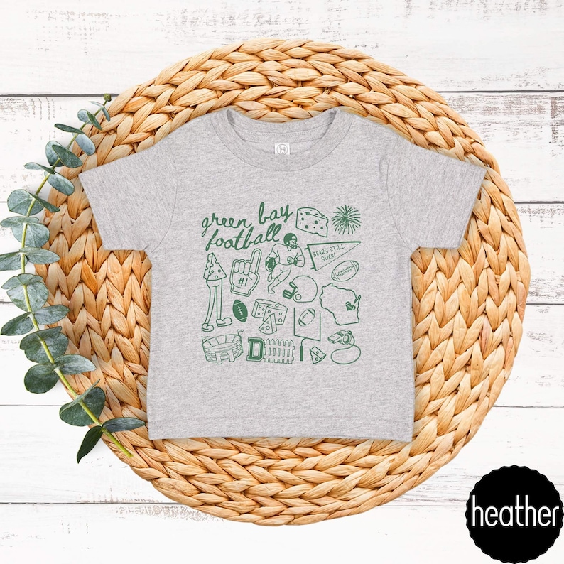 May include: A grey t-shirt with a green doodle design featuring the words "green bay football" and various football-related icons. The shirt is displayed on a woven, circular placemat.