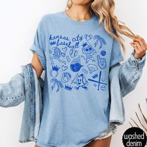 May include: Pale blue t-shirt with a blue graphic design. The design includes the text "Kansas City Baseball" and baseball-themed illustrations. The shirt has short sleeves and a crew neck. The words "washed denim" are printed on a black circle.