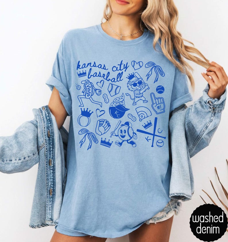 May include: Pale blue t-shirt with a blue graphic design. The design includes the text "Kansas City Baseball" and baseball-themed illustrations. The shirt has short sleeves and a crew neck. The words "washed denim" are printed on a black circle.