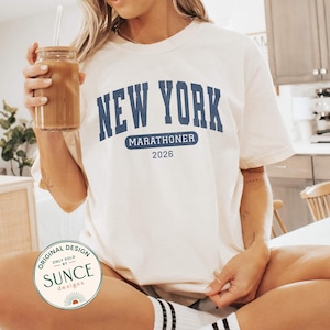 May include: A cream-colored short-sleeve t-shirt featuring the dark blue text NEW YORK in an arched collegiate font, with the word MARATHONER inside a blue rectangle and the year 2026 printed below. The shirt has a crew neckline.