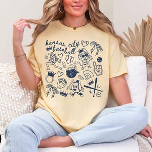 May include: A pale yellow t-shirt with a navy blue graphic design featuring baseball-themed illustrations and the text "Kansas City Baseball". The design includes crowns, hearts, and baseball-related images.