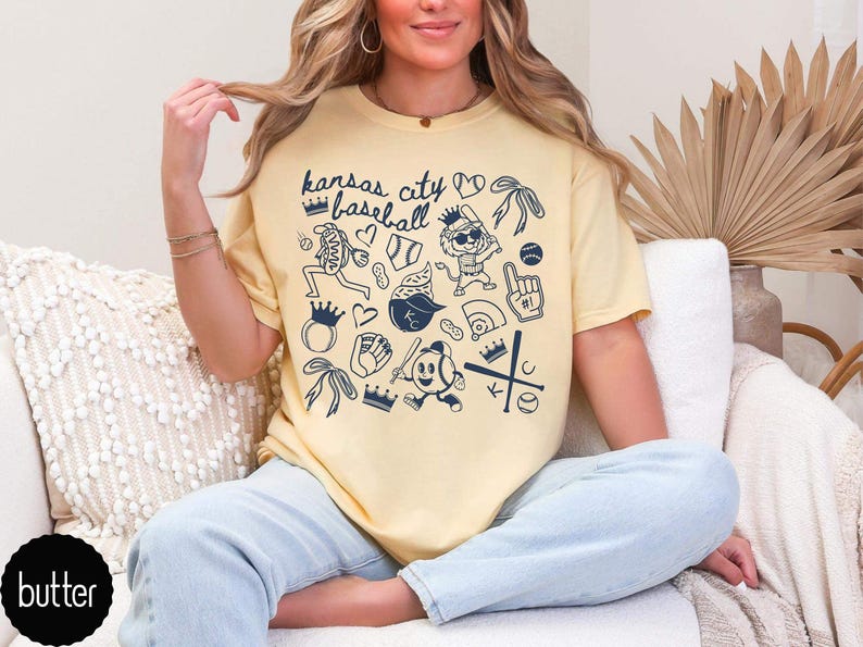 May include: A pale yellow t-shirt with a navy blue graphic design featuring baseball-themed illustrations and the text "Kansas City Baseball". The design includes crowns, hearts, and baseball-related images.