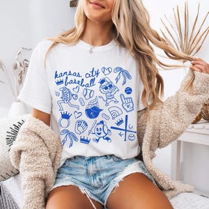 May include: White t-shirt featuring a blue "Kansas City Baseball" design. The graphic includes baseball-related illustrations such as a baseball, glove, and crown. The shirt is paired with denim shorts and a beige cardigan.