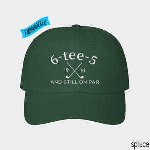 May include: A dark green baseball cap featuring white embroidery. The text reads 6-tee-5, 19 61, and AND STILL ON PAR, accompanied by an illustration of two crossed golf clubs. A blue banner in the upper left corner contains the word EMBROIDERED.