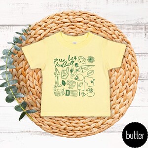 May include: A pale yellow t-shirt with a green graphic design. The design includes the words "Green Bay Football" and football-themed illustrations. The shirt is presented on a woven, circular background.