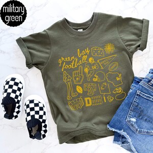 May include: Olive green t-shirt featuring a yellow graphic of football icons and the words "Green Bay Football." The shirt is styled with denim shorts and black and white checkered slip-on shoes. The shirt is labelled "military green."