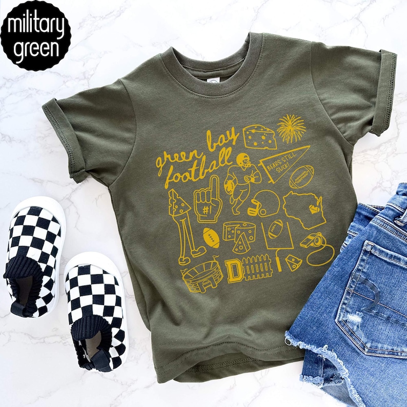 May include: Olive green t-shirt featuring a yellow graphic of football icons and the words "Green Bay Football." The shirt is styled with denim shorts and black and white checkered slip-on shoes. The shirt is labelled "military green."