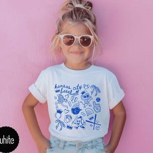 May include: White t-shirt with blue "Kansas City Baseball" graphic. The design includes baseball-themed illustrations like crowns, gloves, and a baseball. The shirt is worn by a young person with sunglasses against a pink background.