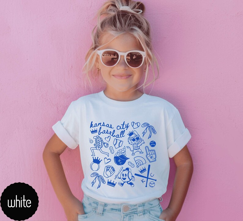 May include: White t-shirt with blue "Kansas City Baseball" graphic. The design includes baseball-themed illustrations like crowns, gloves, and a baseball. The shirt is worn by a young person with sunglasses against a pink background.