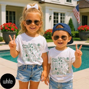 May include: Two children wearing white t-shirts with a green "Green Bay Football" graphic and sunglasses. The shirts feature various football-themed illustrations. They are wearing blue denim shorts and smiling, giving the peace sign.