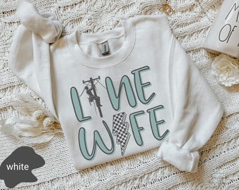 May include: White sweatshirt with the text "Line Wife" in teal blue lettering. A black silhouette of a lineman is climbing a utility pole. A checkered lightning bolt graphic is below the text.