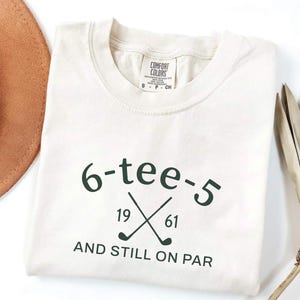 May include: Cream-colored t-shirt with the text "6-tee-5" in dark green, along with crossed golf clubs and the years "19" and "61". The phrase "AND STILL ON PAR" is printed below the graphic.